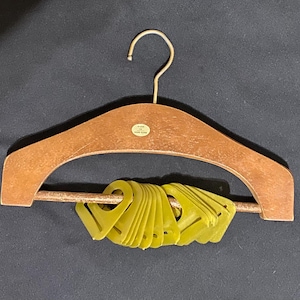 Pode incluir: Um cabide de madeira com um gancho de latão, segurando um feixe de marcadores de tamanho de roupa de plástico amarelo. O cabide tem um design curvo e um pequeno adesivo. O fundo é de cor escura e sólida.