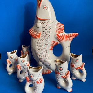 以下が含まれることがあります： 金色の栓が付いた魚の形をした陶器製のピッチャーと、お揃いの小さな魚の形をしたカップ6個のセット。白地にオレンジ色の鱗、ひれ、尾が描かれており、ピッチャーと6個のカップの縁には金色の装飾が施されています。