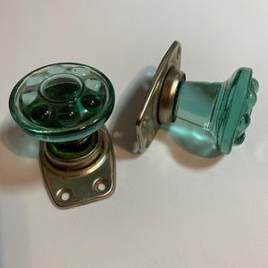 Può includere: Due pomelli per porta in stile vintage realizzati in vetro traslucido verde acqua con un motivo circolare a bolle sulla parte anteriore. Ogni pomello è fissato a una piastra di montaggio metallica rettangolare con due fori per le viti.