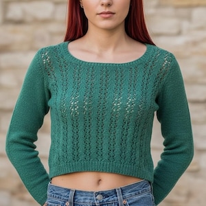 May include: A green, long-sleeved sweater with a round neckline. The sweater has a cropped fit and a textured, open-knit design on the front. The model is wearing blue jeans.