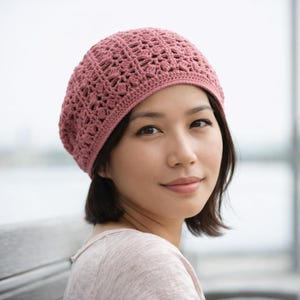 May include: A dusky pink crochet beret with an openwork pattern. The hat is made from a soft, textured yarn and has a rounded crown. The beret is worn by a person with short dark hair.