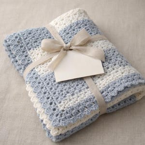 Może przedstawiać: Złożony, szydełkowy koc w odcieniach niebieskiego i białego, zawiązany beżową wstążką i kokardą. Dołączona jest pusta biała etykieta. Koc ma falisty brzeg, dla dziecka.