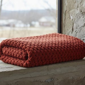 May include: Folded rust-colored chunky knit blanket. The blanket is made of thick yarn and has a textured appearance. It is resting on a stone windowsill, with a blurred outdoor scene visible in the background.