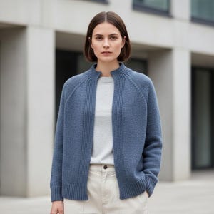 May include: A woman wearing a blue knit cardigan over a light gray shirt and cream-colored pants. The cardigan has a ribbed texture and a stand-up collar. The outfit is suitable for casual or business casual wear.