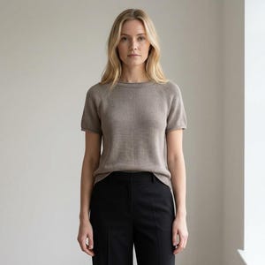 May include: A woman wearing a short-sleeved, light brown knit top and black pants. The top has a crew neck and a ribbed texture. The pants are high-waisted and straight-legged. The background is a plain, light-colored wall with a window on the right.