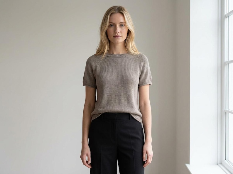 May include: A woman wearing a short-sleeved, light brown knit top and black pants. The top has a crew neck and a ribbed texture. The pants are high-waisted and straight-legged. The background is a plain, light-colored wall with a window on the right.