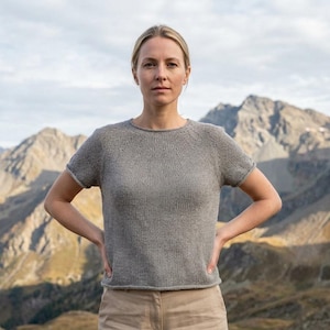 May include: A gray short-sleeved knit top with a crew neck. The top has a simple design and is worn with tan pants. The background features a mountain range under a cloudy sky. The model is standing with their hands on their hips.