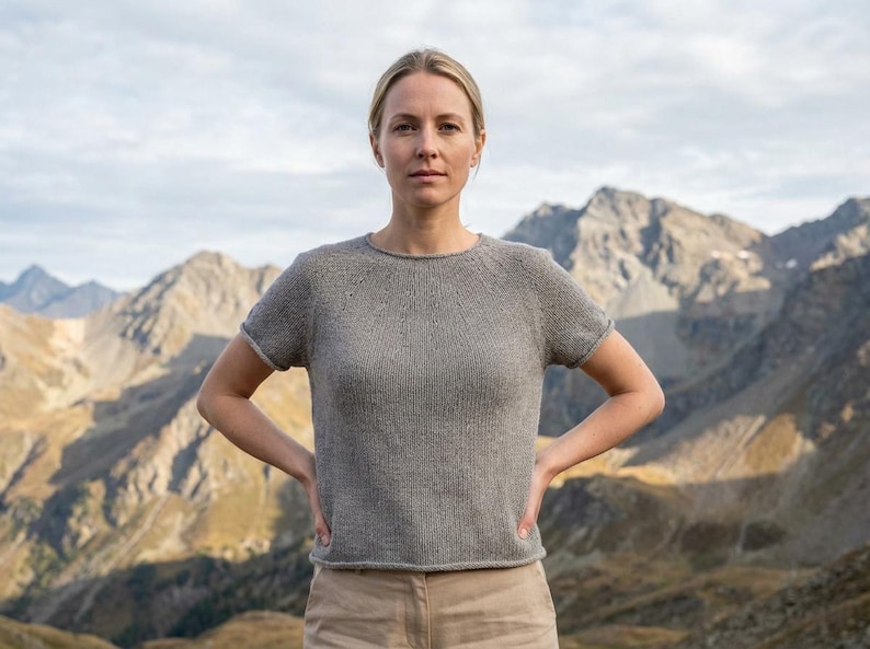 May include: A gray short-sleeved knit top with a crew neck. The top has a simple design and is worn with tan pants. The background features a mountain range under a cloudy sky. The model is standing with their hands on their hips.