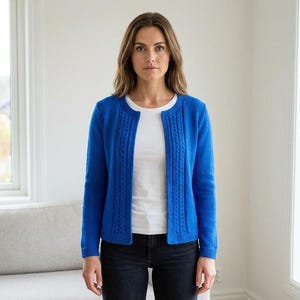 Könnte beinhalten: Eine Frau trägt einen leuchtend blauen Cardigan über einem weißen Oberteil und dunklen Jeans. Der Cardigan hat ein Zopfmuster an den vorderen Kanten. Das Outfit ist schlicht und lässig, geeignet für den Alltag. Der Cardigan ist etwa 100 cm lang.
