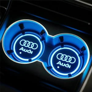 May include: A dual cup holder with a bright blue LED light. The Audi logo is illuminated in white within each circular holder. The cup holder is set in a black interior.