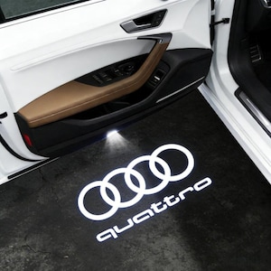 Puede incluir: Una puerta de coche blanca con un interior marrón y negro. La puerta tiene una manija plateada. El suelo muestra el logotipo de Audi y la palabra "Quattro" en blanco, iluminada por una luz.