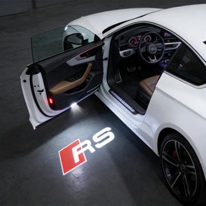 Puede incluir: Coche deportivo blanco con la puerta abierta, que revela un interior beige. La puerta del coche proyecta una luz blanca brillante en el suelo, mostrando las letras "RS" en blanco con un cuadrado rojo.