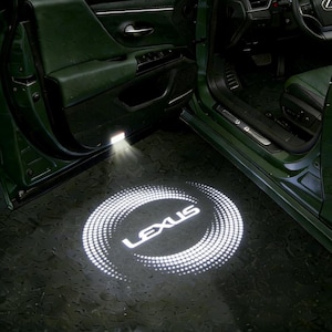 以下が含まれることがあります： 車の内装の詳細。床に白く投影されたレクサスのロゴ。ロゴは点線で囲まれています。車のドアと内装が見え、濃い緑色の配色です。