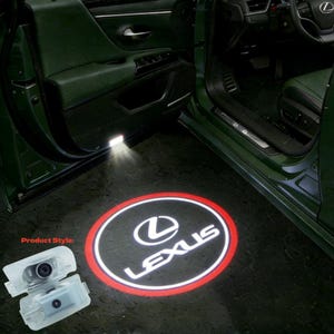 May include: A car door projector displaying the Lexus logo in white with a red outline. The projector is illuminating the ground near the open door of a green car. The product style is visible in the image.
