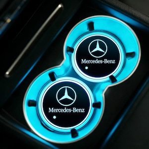 May include: A car cup holder with a bright blue LED light. The cup holder has two circular spaces with the Mercedes-Benz logo in white on a black background. The cup holder is in a black car interior.