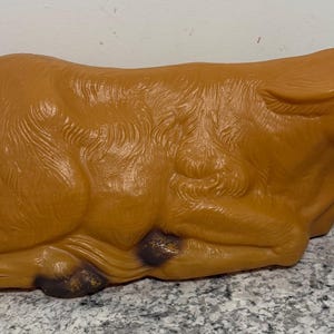 May include: A brown plastic cow figurine in a resting position. The figurine has detailed fur texture and dark brown accents on the hooves and ears. The cow is a decorative item.