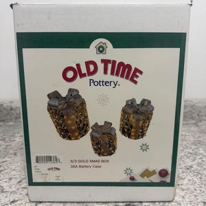 May include: A white cardboard product box for Old Time Pottery featuring three gold-colored, wire-mesh gift box decorations with bows. The box displays the text S/3 GOLD XMAS BOX and 3AA Battery Case, along with a barcode and small snowflake graphics.