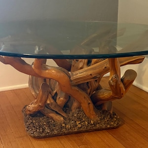 May include: A coffee table featuring a circular clear glass top supported by a base constructed from interconnected, polished light brown tree branches and roots, anchored to a rectangular foundation covered in small, dark brown pebbles.