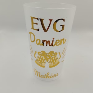 Peut inclure: Un gobelet en plastique blanc givré avec une inscription dorée brillante indiquant EVG, Damien et Mathieu. Le design comprend un motif de deux chopes de bière qui s'entrechoquent avec de la mousse, placé entre les noms en lettres dorées.