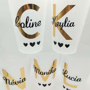 Può includere: Cinque bicchieri di plastica bianca con scritte dorate e nomi in corsivo nero: "boline", "Keylia", "Névia", "Monika" e "Lucia". Ogni bicchiere ha un'iniziale dorata e tre cuori neri.