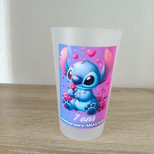 Op de afbeelding: Een matglazen kunststof beker met een kleurrijke afbeelding van een blauw alien-figuur die een roze roos vasthoudt, omringd door roze hartjes en rozen. De tekst onderaan luidt 7 ans Anniversaire ANASTASIA in een wit lettertype.