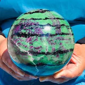 May include: A polished spherical stone featuring a vibrant green base color with prominent horizontal black bands and scattered deep magenta-colored inclusions throughout the surface, creating a natural marbled appearance across the entire sphere.