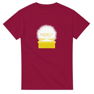 Może przedstawiać: Bordowa koszulka z złotą grafiką "MERCY" nad żółtym prostokątem. Projekt zawiera białe, promieniste tło i tekst na dole. Prosty, odważny design.