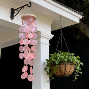 Puede incluir: Un carillón de viento de conchas rosas cuelga de un soporte de metal negro en un porche blanco. El carillón presenta múltiples conchas circulares suspendidas de una parte superior de madera. También se ve una cesta colgante con plantas verdes.