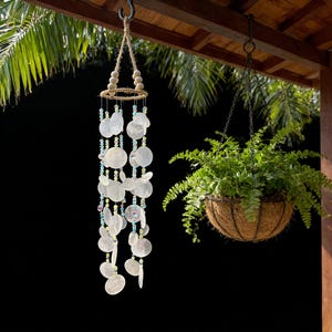 May include: A decorative wind chime featuring white shell discs and colourful beads, suspended from a jute rope and wooden beads. A hanging basket with a green plant is also visible in the image.