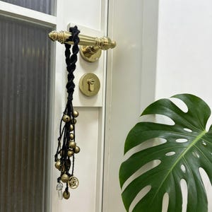 Peut inclure: Un porte-bonheur tressé noir avec des cloches en laiton, un charme de pentagramme et un cristal clair suspendu à une poignée de porte. La poignée et la serrure sont dorées, et la porte est blanche.