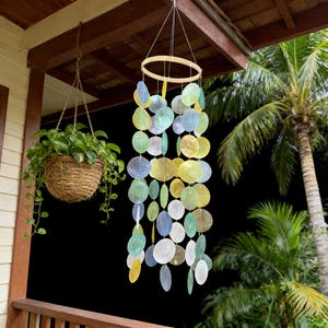 Peut inclure: Un carillon éolien suspendu, composé d'un anneau en bois et de rangées de disques de coquillages irisés aux couleurs jaune, vert, bleu et blanc. Le carillon est suspendu à un avant-toit. Un panier de plantes suspendu est visible en arrière-plan.