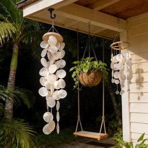 Puede incluir: Escena al aire libre con decoraciones colgantes. Un carillón de viento hecho de conchas blancas y redondas cuelga de un gancho. También se ve una cesta colgante con plantas verdes y un columpio de madera. La escena está al aire libre.