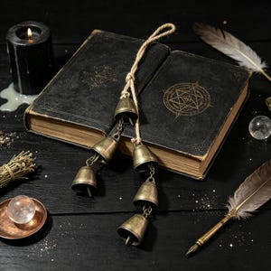 May include: A set of five antique-style brass bells suspended from a natural jute twine rope, resting on a black hardcover book featuring a gold embossed star symbol. The scene includes a black candle, a clear crystal sphere, and a quill pen.