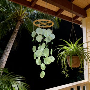 May include: A hanging wind chime with light green, disc-shaped elements suspended from a wooden ring. The chime is hanging from a porch ceiling, with a potted plant to the right and a palm tree in the background.