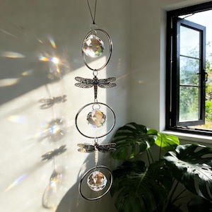 May include: A decorative suncatcher featuring three crystal spheres within silver rings, with bronze dragonfly charms. The suncatcher hangs in front of a window, creating a rainbow effect from the sunlight. The spheres and rings are suspended from a black cord.