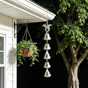 May include: A silver wind chime with five bells hangs from a roof. The top of the chime features a dragon design. A hanging basket with green plants is also visible, suspended from a black metal bracket.