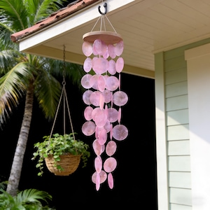 Könnte beinhalten: Ein rosa Capiz-Muschel-Windspiel, das an einer Holzscheibe hängt. Das Windspiel besteht aus mehreren Strängen durchscheinender, runder und ovaler Muscheln. Das Windspiel hängt an einem schwarzen Haken an einer Veranda, mit einem hängenden Pflanzenkorb im Hintergrund.