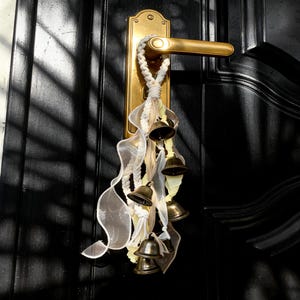 May include: A decorative door hanging featuring small bronze bells, cream-colored rope, and sheer ribbons in cream and pale yellow. The bells are suspended from a rope tied around a gold-colored door handle and plate on a black door.