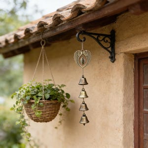 May include: A hanging wind chime with four bronze-colored bells and a decorative heart-shaped top with wing details. The chime hangs from a black metal bracket on a stucco wall. A hanging woven basket with green plants is also visible.