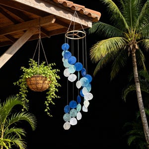 May include: A hanging wind chime with blue and white shell-shaped accents. The chime is suspended from a gold-colored ring. A hanging basket with green plants is to the left. Palm trees and a dark background complete the scene.