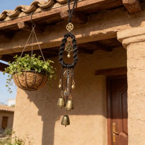Puede incluir: Un colgante de pared de macramé negro con un pentagrama, un nudo celta y un dije de llave. El colgante también presenta pequeñas campanas y cuentas de color dorado. El fondo incluye una cesta colgante de vegetación y una pared de estuco.