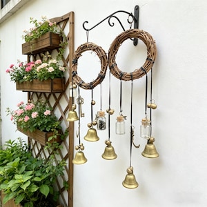 Può includere: Due ghirlande circolari di vite appese a una staffa di metallo nero. Ogni ghirlanda ha nastri neri con piccole campane di ottone, bottiglie di vetro e ciondoli decorativi. Lo sfondo presenta un muro bianco e fioriere di legno con fiori rosa.