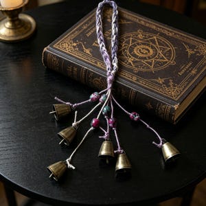 May include: A handmade wind chime with small brass bells and decorative beads in purple, pink, and green. The bells hang from a braided cord, placed on a vintage book with gold lettering.