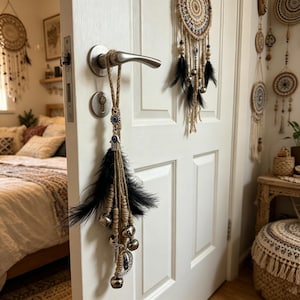 Puede incluir: Un adorno decorativo para puerta con cordón de yute, plumas negras, campanas plateadas y un amuleto de ojo turco. El adorno cuelga de una manija de puerta plateada. Se ven atrapasueños y decoración bohemia en el fondo.