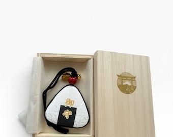 May include: A triangular omamori charm shaped like an onigiri rice ball, featuring a white textured surface, a black strip, and gold Japanese characters. It includes a black cord, a red bead, and a small gold bell, presented inside a light wood box.
