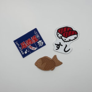 May include: Three embroidered patches: a rectangular blue patch with red Japanese text and white graphics, a white patch shaped like sushi with red and black details and Japanese text, and a brown fish-shaped patch with textured scale details.