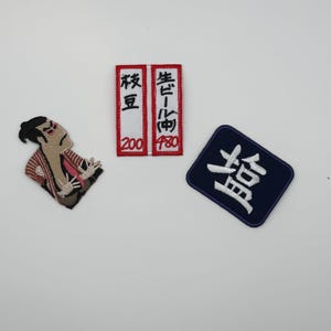 以下が含まれることがあります： 3つの刺繍ワッペン。赤と白の柄の服を着た人物のワッペン、赤い縁取りで黒い日本語の文字と赤い数字が書かれた白い長方形のワッペン、白い日本語の文字が書かれた濃い青色の長方形のワッペン。
