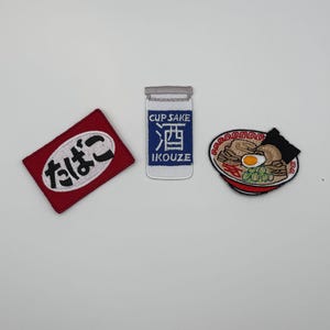 Peut inclure: Trois écussons brodés comprenant un écusson rectangulaire rouge avec du texte japonais blanc, un écusson rectangulaire bleu et blanc marqué CUP SAKE IKOUZE avec un caractère japonais, et un écusson circulaire représentant un bol de ramen.