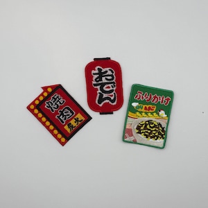 以下が含まれることがあります： 日本をテーマにした3つの刺繍ワッペン。1つは黄色いドットと黒い文字が入った赤い長方形の看板、1つは黒い文字が入った赤い提灯の形、もう1つは食べ物のボウルと日本語の文字が描かれた緑色の長方形のパッケージです。