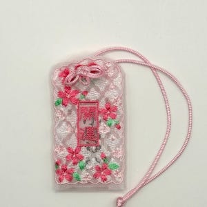 以下が含まれることがあります： 白いレースが重なった長方形のピンク色のお守り。ピンクの桜の花と緑の葉が刺繍されています。中央には濃い赤色の漢字が書かれたピンクの長方形の布地があり、上部にはピンクの紐が付いています。